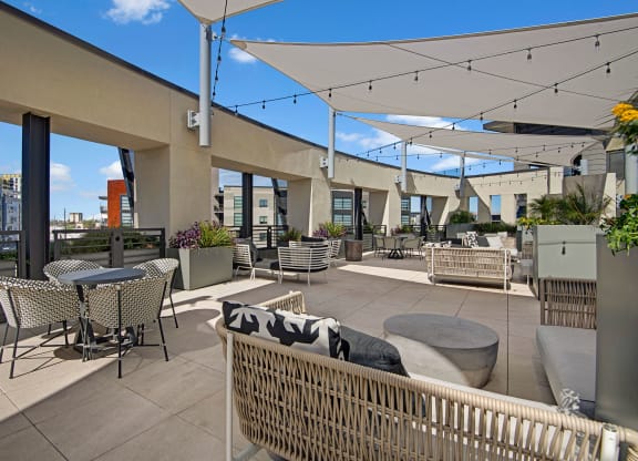Rooftop terrace here at Roosevelt Row Apartments with modern seating, tables, and planters under white shade sails. String lights add ambiance. City skyline visible under a clear blue sky.