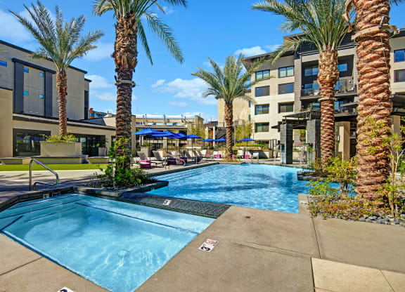 Modern outdoor pool area here at Roosevelt Row Apartments with palm trees, a jacuzzi, and poolside lounge chairs under umbrellas. Sunny day with a hotel building in the background.