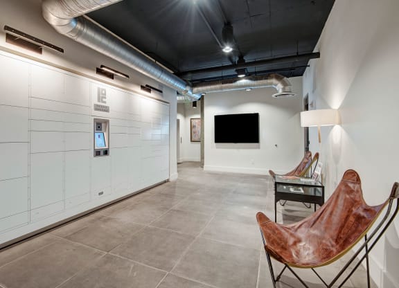 A modern lobby here at Roosevelt Row Apartments featuring white parcel lockers with a digital screen on the left. Two brown leather butterfly chairs and a lamp on the right, all under exposed ductwork.