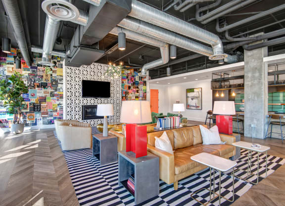 Modern lounge here at Roosevelt Row Apartments with eclectic decor featuring a tan leather sofa, colorful wall collage, striped rug, and exposed ducts. Bright, vibrant, and inviting atmosphere.