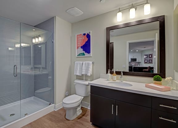 Modern bathroom here at Roosevelt Row Apartments with glass shower, dark wood vanity, and large mirror. Bright lighting, white towels, and art poster create a clean, stylish feel.