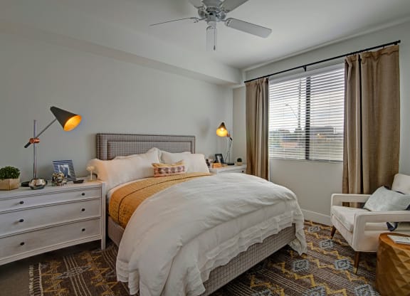 Cozy bedroom here at Roosevelt Row Apartments with a large bed, white and mustard bedding, modern lamps, and a patterned rug. Natural light filters through blinds, creating a warm, inviting atmosphere.