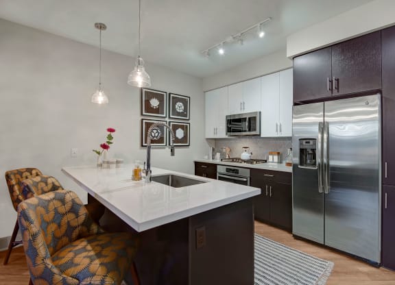 Modern kitchen here at Roosevelt Row Apartments with dark wood cabinets, white countertops, and stainless steel appliances. Two patterned chairs at the island, pendant lights, and floral decor add elegance.