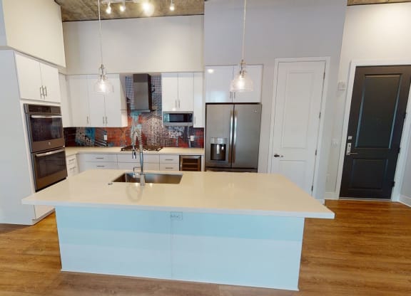 Modern kitchen here at Roosevelt Row Apartments with wooden floors, a large island with a sink, pendant lights, white cabinets, and colorful mosaic backsplash create a sleek look.