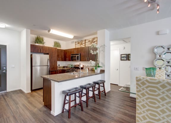 Modern kitchen here at Artisan at Main Street Metro Apartments with dark wood cabinets, stainless steel appliances, and a granite island featuring bar stools. Bright, airy, with decorative plants.