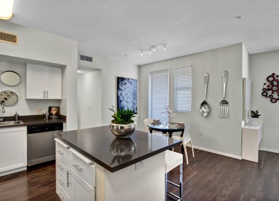 Modern kitchen and dining area here at Artisan at Main Street Metro Apartments with dark wood floors, white cabinets, and a gray countertop island. Wall decor includes oversized silver utensils.