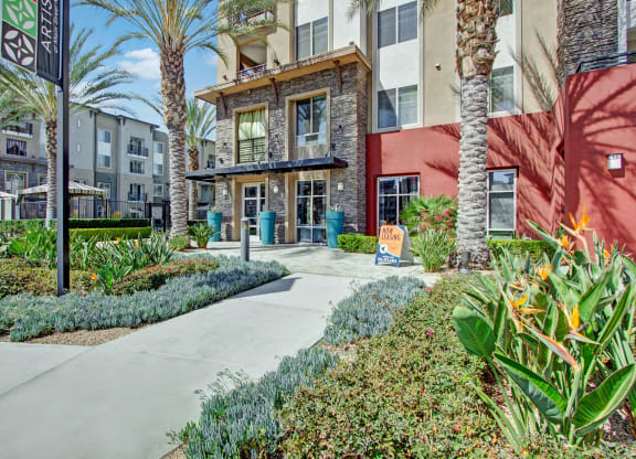Exterior of our modern Artisan at Main Street Metro apartment complex with red accents, palm trees, and lush landscaping. A "Now Leasing" sign is displayed near the entrance.