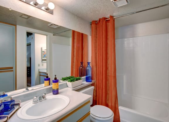 Small bathroom here at Castlepark Apartment Homes with a white vanity and sink, orange shower curtain, and white tiles. Blue glass bottles and a green potted plant add color. Clean and inviting.