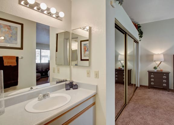 Bright bathroom here at Castlepark Apartment Homes with a lit vanity mirror, geometric art, and towels. Adjacent bedroom has mirrored closet doors, a bedside lamp, and dresser. Cozy and inviting atmosphere.