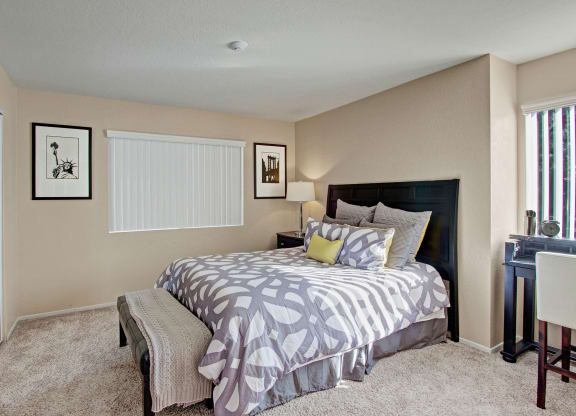 Cozy bedroom here at Castlepark Apartment Homes with a modern design. Features a bed with patterned gray and white bedding, a bedside lamp, abstract framed art, and a small desk by the window.