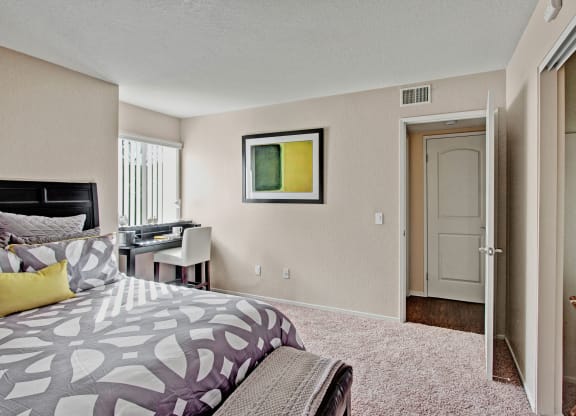 Bright bedroom here at Castlepark Apartment Homes with a modern, gray and white patterned bedspread, yellow accent pillow. A window desk and chair on the left; open door to the right.