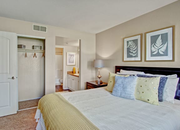 Cozy bedroom here at Castlepark Apartment Homes with a neatly made bed, yellow and gray pillows, fern artwork, open closet, and bathroom view. Warm, inviting ambiance.