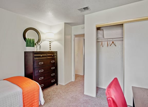 Bedroom here at Castlepark Apartment Homes with white walls, a bed with an orange cover, a tall dark dresser with a lamp and decor on top, an open closet, and a red chair next to a desk.