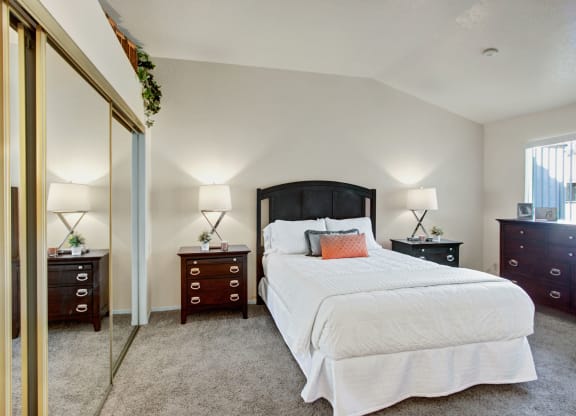 Cozy bedroom here at Castlepark Apartment Homes with a plush carpet features a central bed with white bedding, wooden headboard, two bedside tables with lamps, and mirrored closet doors.