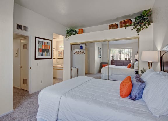 Bedroom here at Castlepark Apartment Homes with a cozy feel, showcasing a neatly made bed with white linens and orange accent pillows. Large mirror, decorative plants, and modern artwork adorn the walls. Soft lighting adds a warm ambiance.