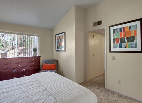 Bedroom here at Castlepark Apartment Homes with cream walls, featuring a bed with a white quilt, a dark wood dresser by a window, and colorful abstract wall art. Cozy and serene ambiance.