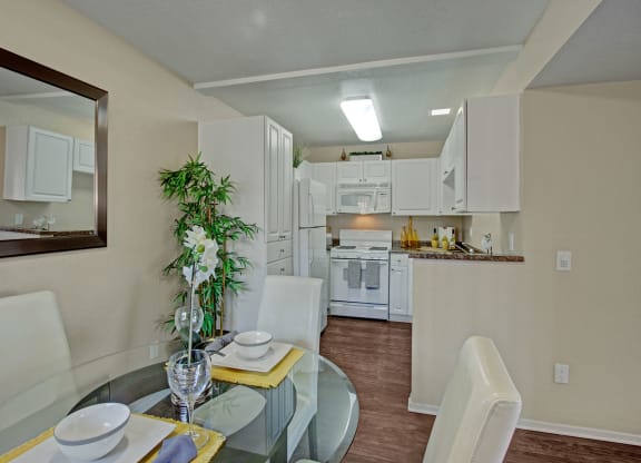 Modern kitchen and dining area here at Castlepark Apartment Homes with white cabinetry, a glass table, and leather chairs. A potted plant adds greenery; soft lighting creates a cozy atmosphere.