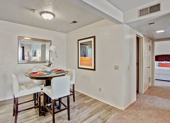 Cozy dining area here at Castlepark Apartment Homes with a round glass table, white chairs, and an orchid centerpiece. Mirror on one wall; adjacent to a hallway leading to a bedroom. Warm, inviting tone.