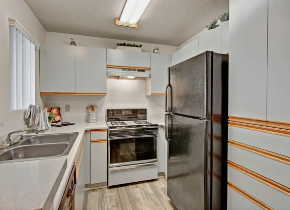 Compact kitchen here at Castlepark Apartment Homes with white cabinets, a black refrigerator, gas stove, and stainless steel sink. Bright fluorescent lighting and a tidy, modern appearance.