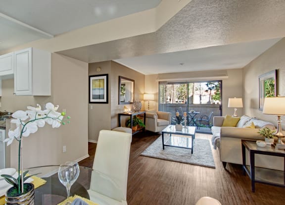Bright, modern open-plan living room and kitchen here at Castlepark Apartment Homes. Features neutral tones, white cabinets, cozy sofas, large window with a view, and elegant decor.