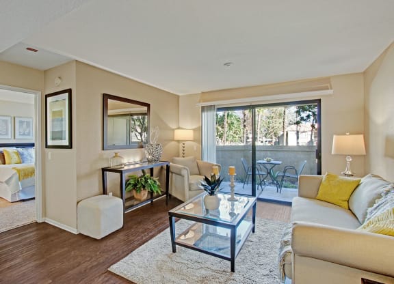 Chic living room here at Castlepark Apartment Homes with beige walls and dark wood floor. Cozy sofa with yellow pillows, glass table, mirror, and indoor plants. Sunny patio view through sliding doors.