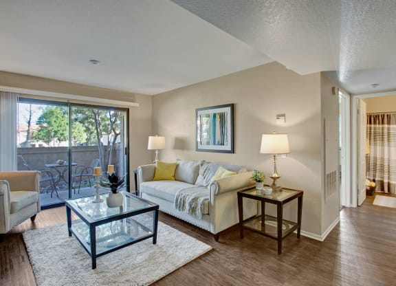 Contemporary living room here at Castlepark Apartment Homes with a white sofa, yellow pillows, and a glass coffee table. Large window shows a patio. Warm lighting creates a cozy ambiance.