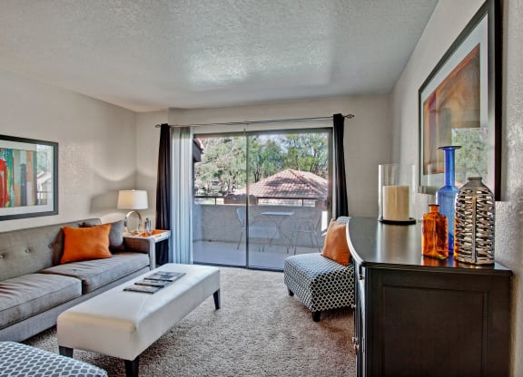 Modern living room here at Castlepark Apartment Homes with a gray couch adorned with orange pillows, complemented by a white ottoman. Large glass doors open to a balcony with trees beyond.