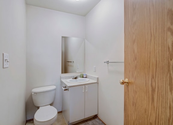 Small, simple bathroom here at Carrington Place Apartment Homes with white walls and neutral tones. Features a toilet, a small mirror above a sink, and wooden door, conveying a minimalistic tone.