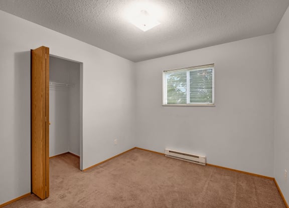 Empty bedroom here at Carrington Place Apartment Homes with beige carpet and light gray walls. Open closet with a wooden door on the left, a small window with blinds on the right. Cozy and minimalistic.