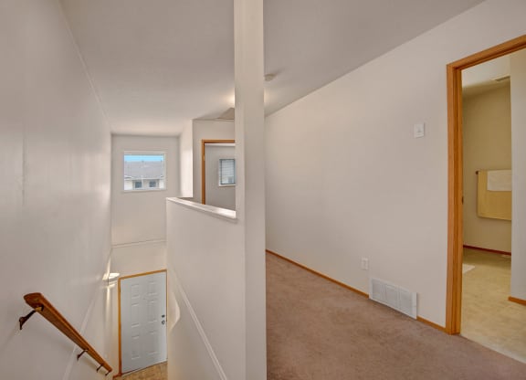 View of a simple, light-filled hallway here at Carrington Place Apartment Homes with white walls, a beige carpet, and a wooden railing on the stairs. A window with a view of rooftops is at the end, and a bathroom door with a towel is visible to the right.