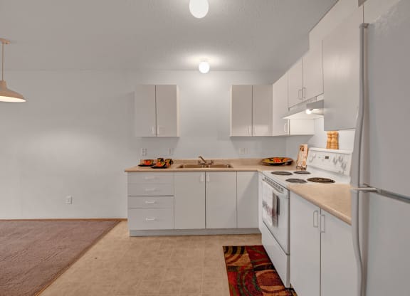 Simple kitchen here at Carrington Place Apartment Homes with white cabinets, beige countertops, and white appliances, including a stove and fridge. A colorful rug and bowls add warmth.