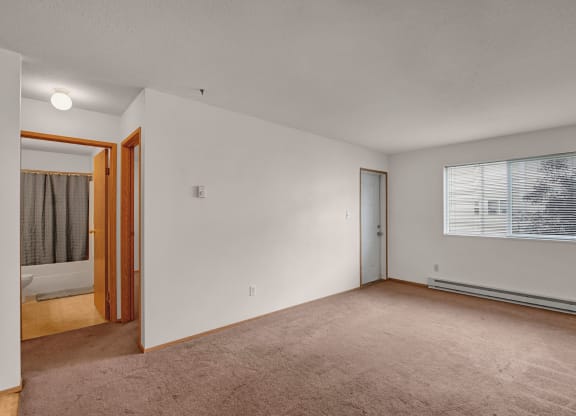 Empty bedroom here at Carrington Place Apartment Homes with beige carpet and white walls. A window with blinds lets in soft daylight. Open door reveals a bathroom with a shower curtain. Minimalist tone.