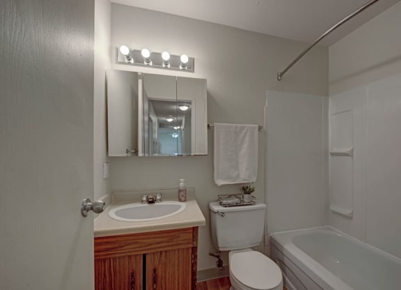 A small bathroom here at Cedar Gardens Apartment Homes with a wooden vanity, oval sink, and mirror cabinet. It includes a toilet, a white towel hanging, and a tub with a shower curtain rod. Bright lighting creates a clean and cozy atmosphere.