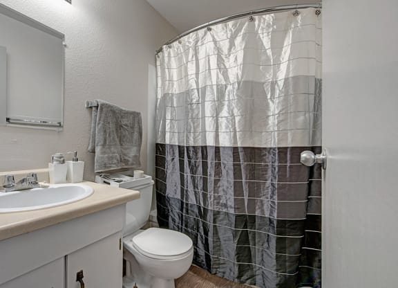 Small bathroom here at Cedar Gardens Apartment Homes with a beige countertop, white sink, and silver faucet. Gray towel hangs above the toilet. Black-and-white shower curtain adds contrast.