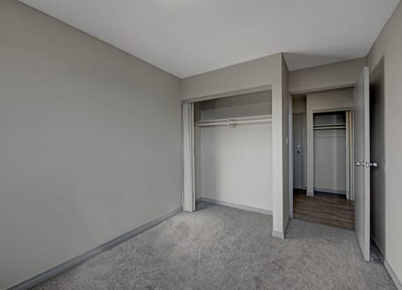 Empty bedroom here at Cedar Gardens Apartment Homes with gray walls and carpet, featuring an open closet with hanging rods. Doorway leads to hardwood floor hallway, creating a minimalist, neutral tone.