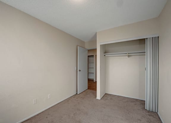 Empty beige room here at Cedar Gardens Apartment Homes with a carpeted floor, partially open closet with sliding door, and an open door leading to a hallway with hardwood flooring. Minimalist and neutral.