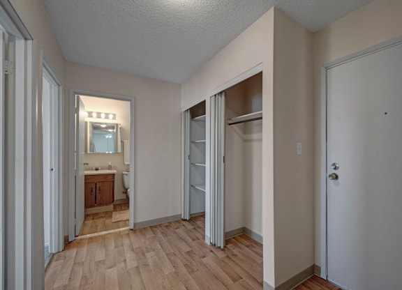 Minimalist hallway here at Cedar Gardens Apartment Homes with light wood flooring. Open closet with sliding doors on the right. Bathroom with mirror and lights visible in the background.