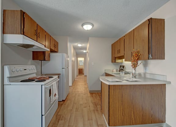 Narrow kitchen here at Cedar Gardens Apartment Homes with wooden cabinets and light countertops. Appliances include a stove and refrigerator. A small vase with flowers adds warmth.