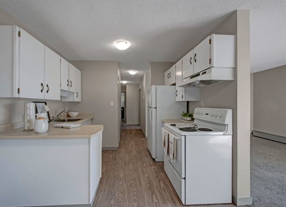 A modern, narrow kitchen here at Cedar Gardens Apartment Homes with white cabinets, an electric stove, and a fridge. Light wood flooring and beige walls create a clean, neutral tone.