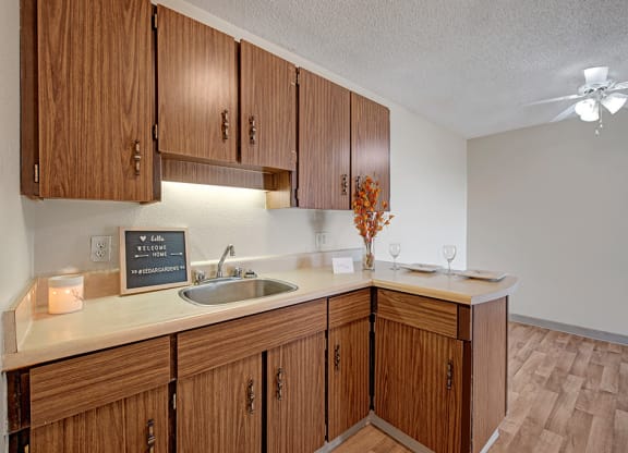 Cozy kitchen here at Cedar Gardens Apartment Homes with wooden cabinets, a small sink, and a lighted backsplash. A welcoming sign, candle, and two place settings add warmth.