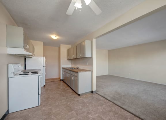 Simple kitchen here at Cedar Gardens Apartment Homes with white appliances, including a stove and refrigerator. Gray cabinets line the walls. Adjacent is a carpeted living room, creating an open and neutral space.