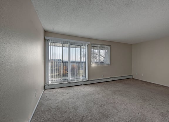 Spacious, empty living room here at Cedar Gardens Apartment Homes with beige walls and carpet. Large sliding glass doors and window with vertical blinds reveal a view of trees and grass outside. Quiet and serene.