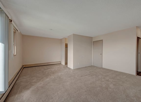 An empty room here at Cedar Gardens Apartment Homes with beige walls and carpet, featuring a closed door, small window on the left, and open closet. Soft lighting creates a calm atmosphere.