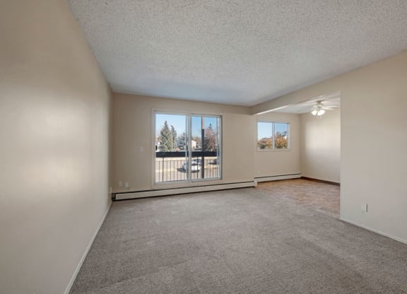 Spacious, empty living room here at Cedar Gardens Apartment Homes with beige walls and carpet, large window showing trees outside. Adjacent dining area with a ceiling fan and tiled floor.
