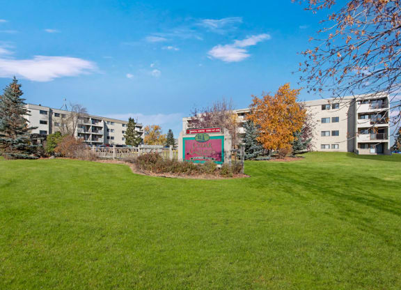 Expansive grassy lawn with a sign for “Cedar Gardens” centered between two beige apartment buildings. Bright autumn trees under a clear blue sky.