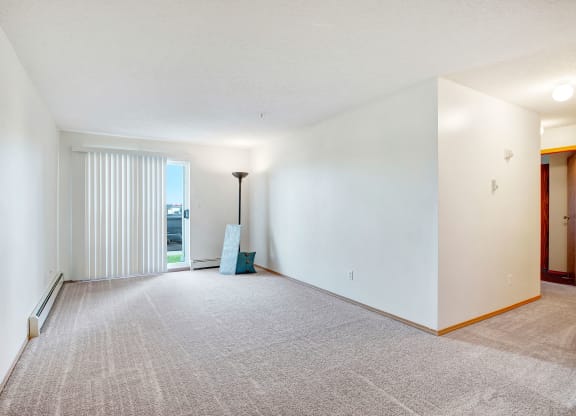 Spacious empty room here at Edgewood Estates Apartment Homes with carpeted floor, white walls, and vertical blinds covering a glass door leading outside. A floor lamp and a cushion add minimal decor.