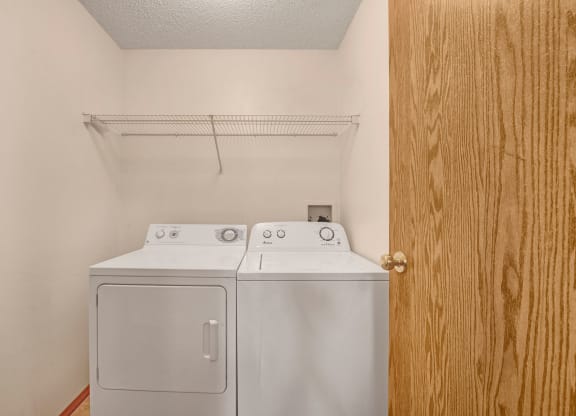 Laundry room here at Edgewood Estates Apartment Homes with a white washer and dryer side by side under a wire shelf on a beige wall. A wooden door is partially open on the right.