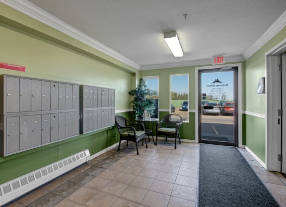 Edgewood Estates Apartment Homes lobby with pale green walls, a wall of mailboxes, and wicker chairs around a small table featuring a plant. A glass door leads outside.