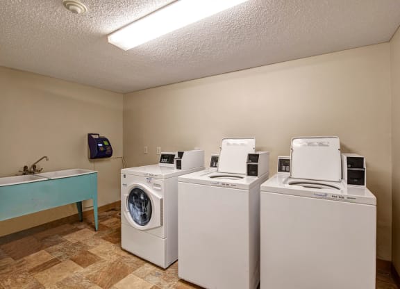 Laundry room here at Lakewood Heights Apartment Homes with two white top-load washing machines and a front-load dryer. A utility sink with a faucet is on the left. Beige walls and tiled floor.