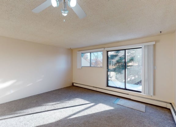 Empty room here at Lakewood Heights Apartment Homes with beige walls, carpeted floor, and ceiling fan. Large sliding glass doors and a window let in sunlight, casting shadows across the room.